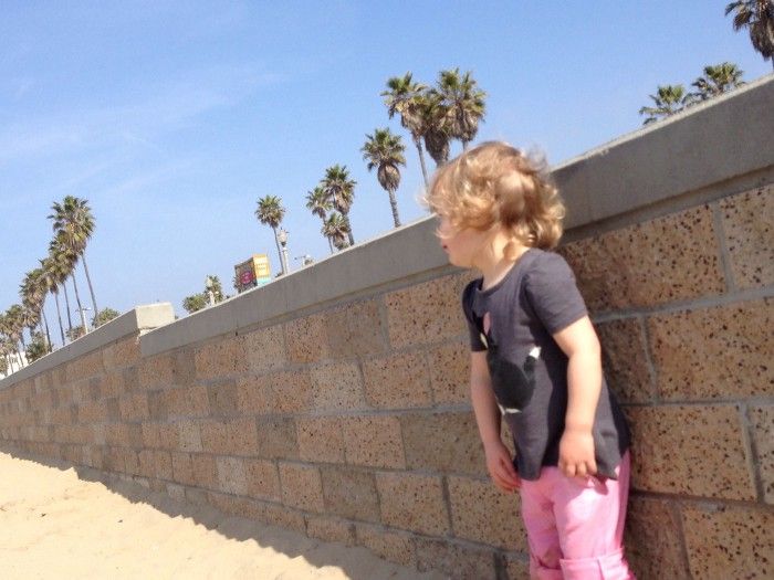 A young girl stands against a seawall at a California beach, with palm trees in the background.