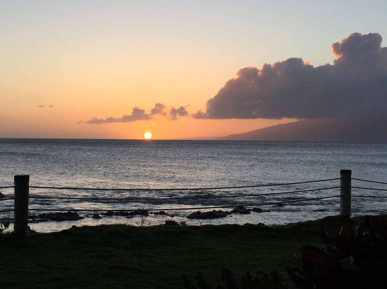 The sun sets over ocean off of Maui, Hawai'i; a small backyard and rope fence is in the foreground.